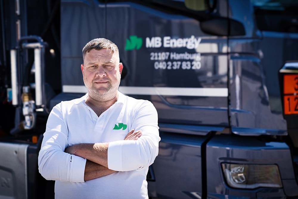 Mitarbeiter in weißem Poloshirt mit verschränkten Armen vor einem MB-Energy-Tanklastwagen.